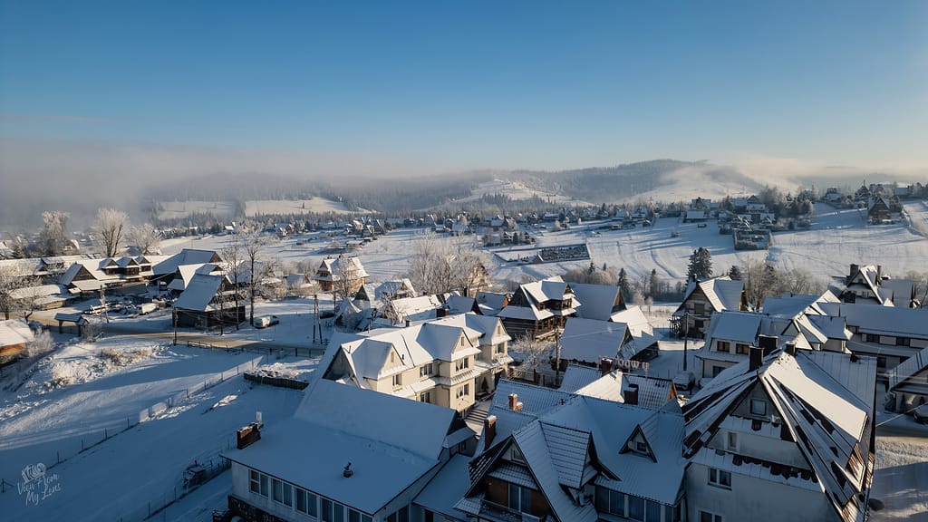 Thrilling Epic Christmas 2025 Murzasichle Tatra Adventures: Aerial view of snow-covered village with triangular-roofed chalets nestled under blue skies, misty hills and frosted forests beyond.